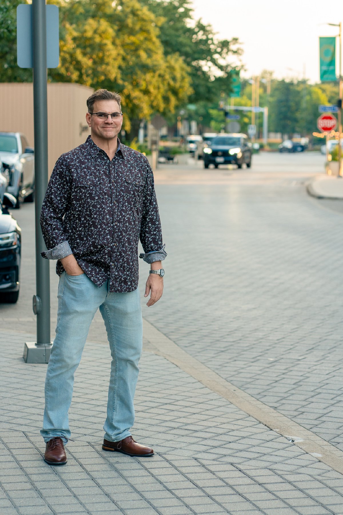 Full-length portrait on a Legacy West street at golden hour, floral shirt with rolled cuffs, light jeans, and brown shoes