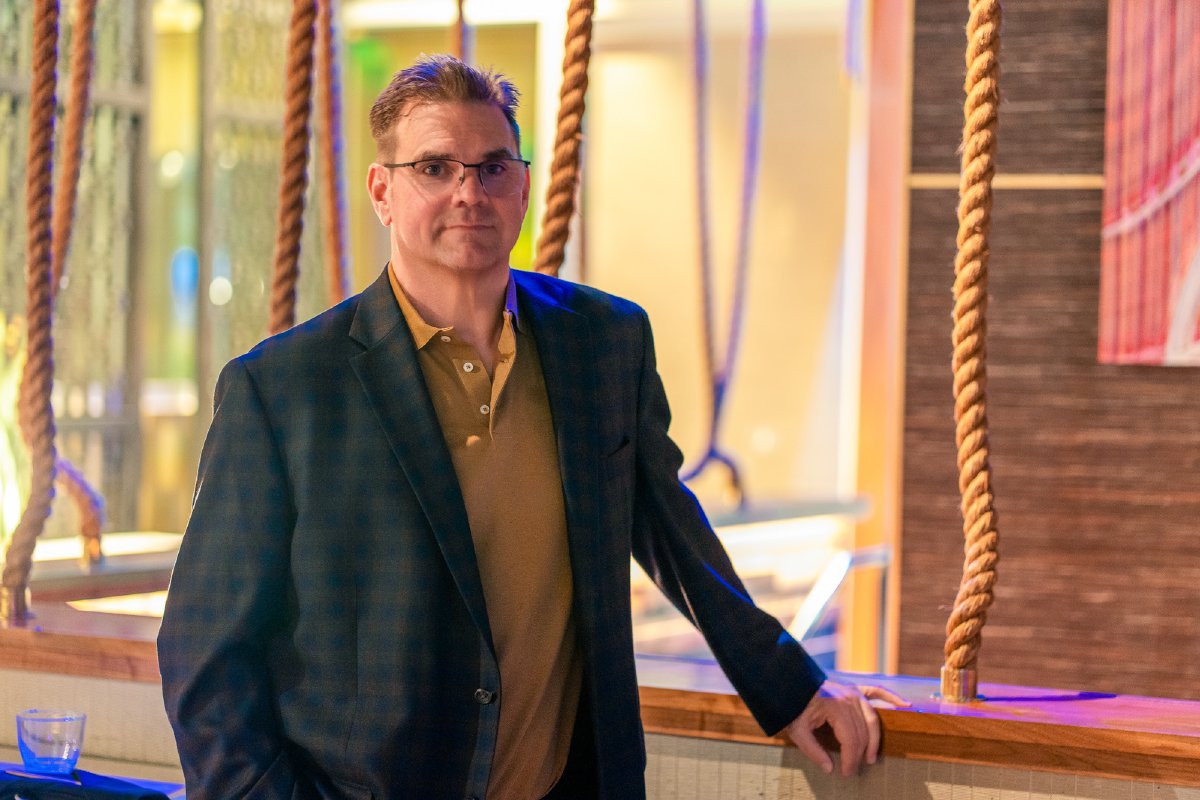 Man in a dark plaid blazer over a tan polo, leaning on a wooden rail between thick hanging ropes