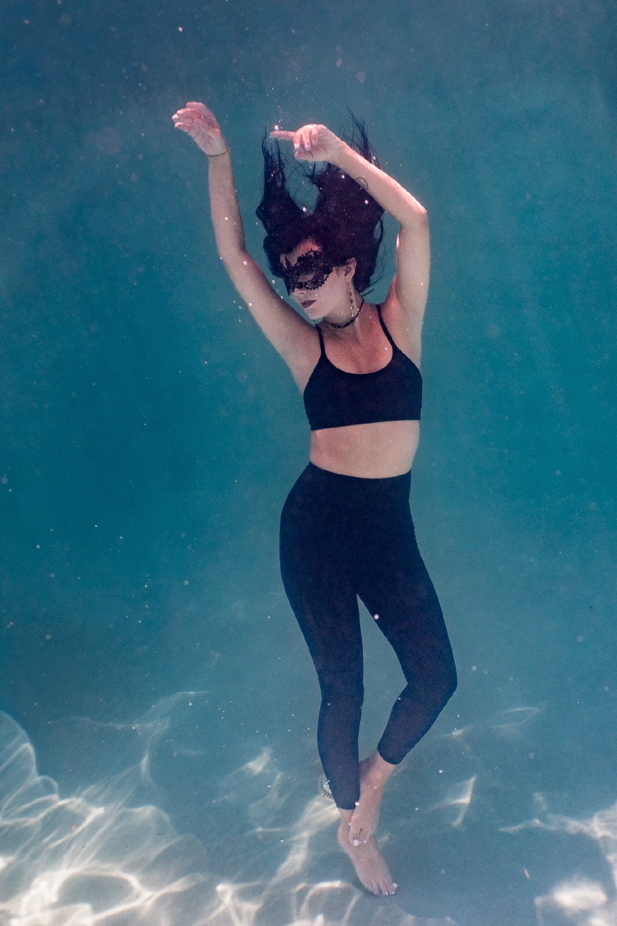 Underwater portrait of a model in a black lace masquerade mask, hair fanned out in deep blue water
