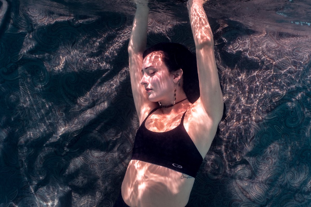Underwater portrait lit by sunlight caustics, arms overhead against a paisley fabric backdrop