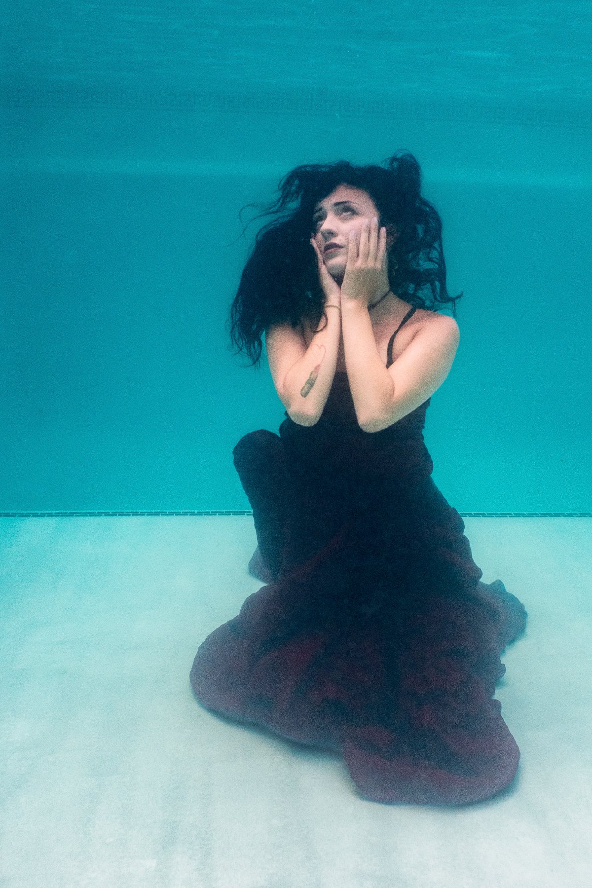 Model seated on the pool floor in a long dark dress, looking up through the water