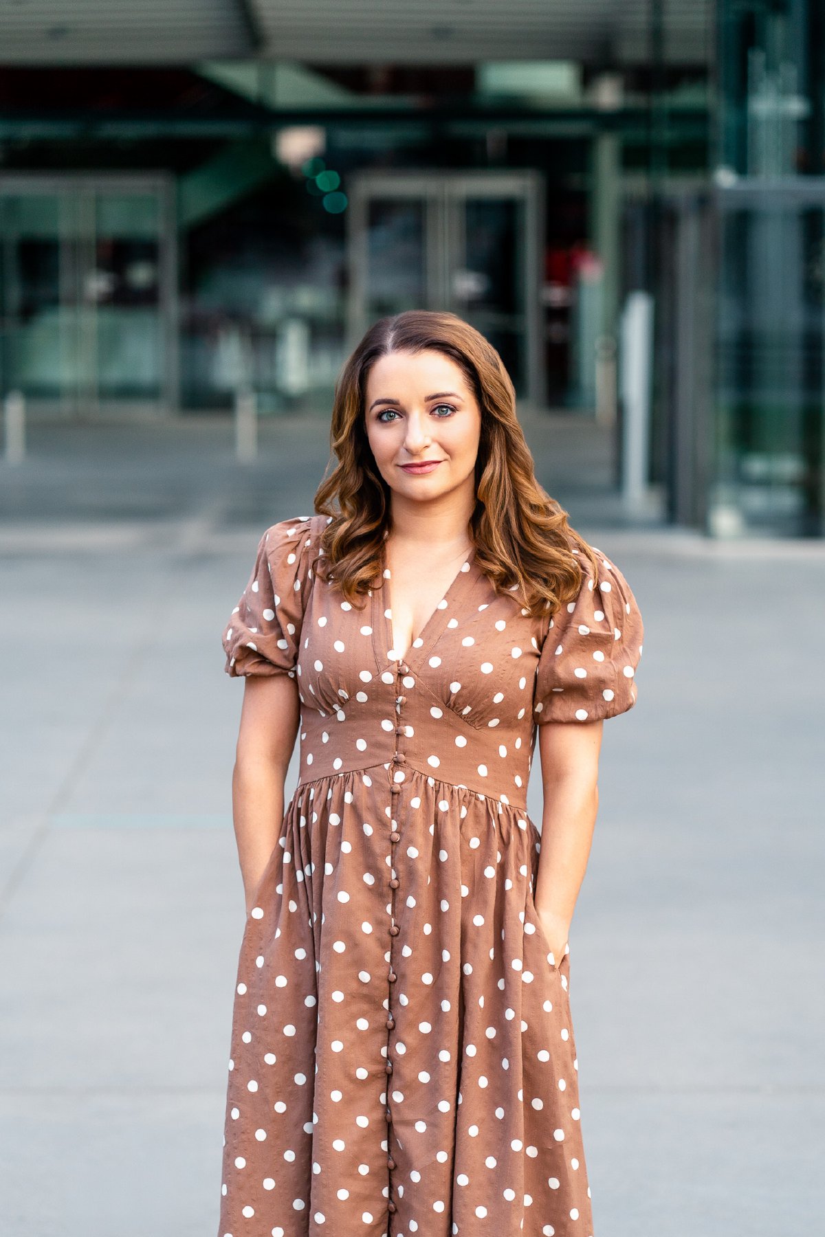 Soraya Mafi in a brown polka dot dress with a soft smile, standing in front of a glass building