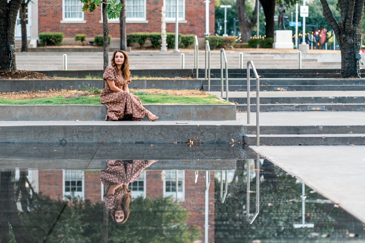 Sitting on a concrete ledge above a still reflecting pool that mirrors her, red brick building behind