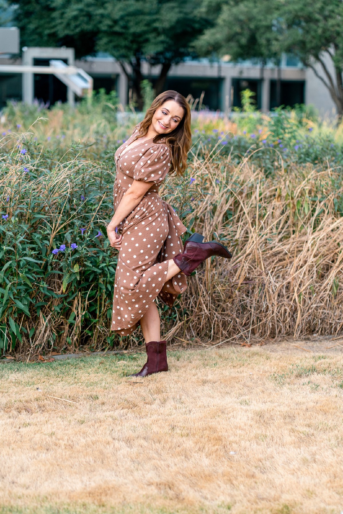 Playful kick in burgundy cowboy boots, laughing in front of tall grass and purple wildflowers