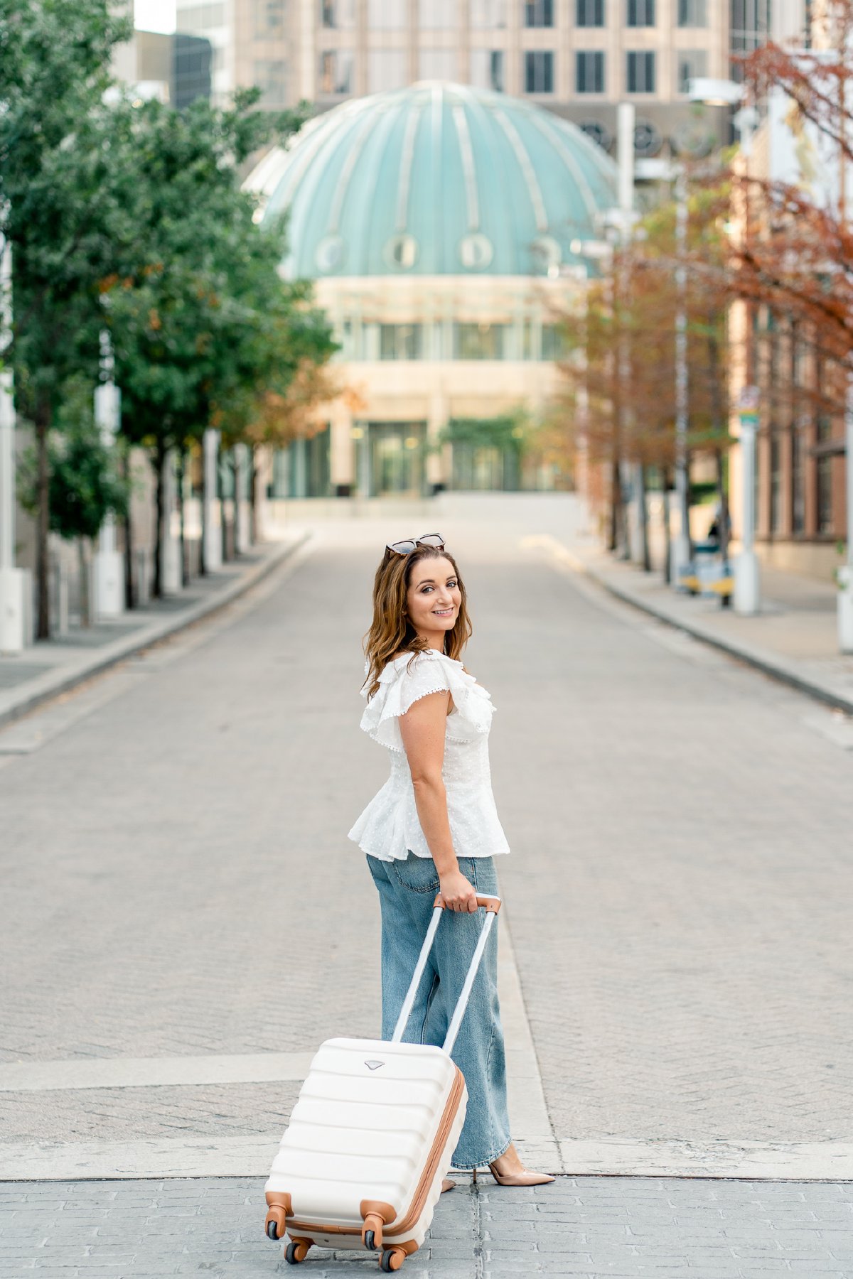 Rolling a white carry-on suitcase down an empty street, glancing back over her shoulder, green dome at the end of the block