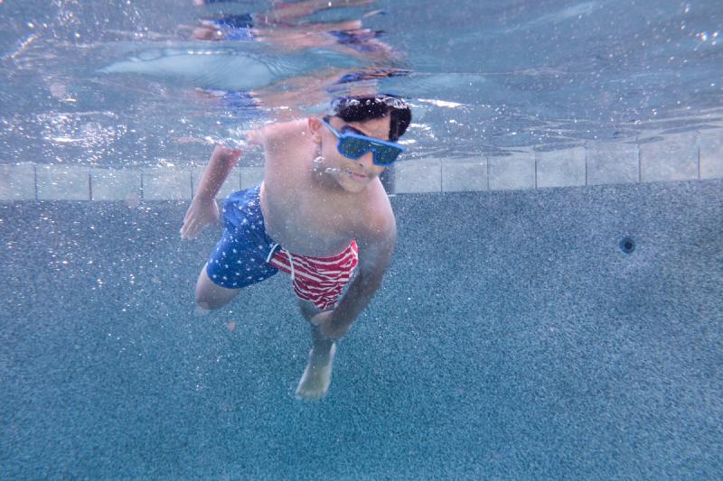 Kid swimming underwater in American flag swim trunks and blue goggles